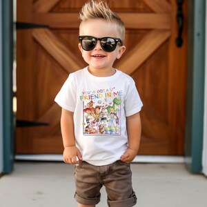 Op de afbeelding: Een wit T-shirt met een kleurrijke afbeelding van personages uit de film "Toy Story" en de tekst "You've Got a Friend in Me". Het shirt wordt gedragen door een jong kind met een zonnebril en een bruine korte broek.