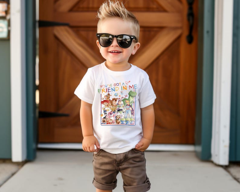 Op de afbeelding: Een wit T-shirt met een kleurrijke afbeelding van personages uit de film "Toy Story" en de tekst "You've Got a Friend in Me". Het shirt wordt gedragen door een jong kind met een zonnebril en een bruine korte broek.