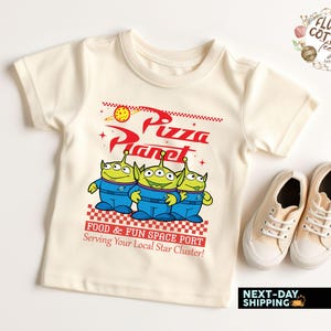 May include: Cream-colored t-shirt featuring the Pizza Planet logo and three green alien characters in blue uniforms. The shirt includes the text "FOOD & FUN SPACE PORT Serving Your Local Star Cluster!" and is paired with beige sneakers.