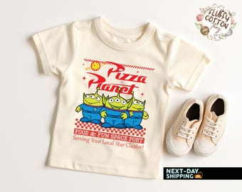 Puede incluir: Camiseta color crema con el logo de Pizza Planet y tres personajes alienígenas verdes con uniformes azules. La camiseta incluye el texto "FOOD & FUN SPACE PORT Serving Your Local Star Cluster!". Se muestra con zapatillas beige.