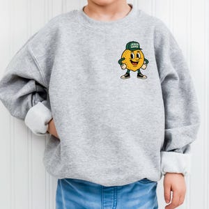 K&ouml;nnte beinhalten: Hellgraues Sweatshirt mit einem Cartoon-Zitronen-Charakter, der eine gr&uuml;ne Kappe mit dem Text "JESUS SAVES" tr&auml;gt. Die Zitrone hat ein l&auml;chelndes Gesicht und tr&auml;gt schwarze Turnschuhe. Das Sweatshirt hat einen Rundhalsausschnitt und lange &Auml;rmel.