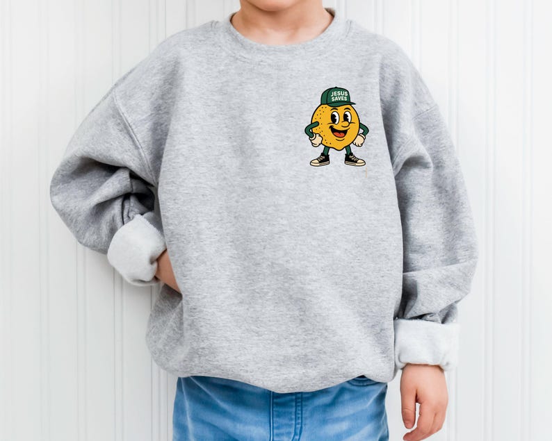 K&ouml;nnte beinhalten: Hellgraues Sweatshirt mit einem Cartoon-Zitronen-Charakter, der eine gr&uuml;ne Kappe mit dem Text "JESUS SAVES" tr&auml;gt. Die Zitrone hat ein l&auml;chelndes Gesicht und tr&auml;gt schwarze Turnschuhe. Das Sweatshirt hat einen Rundhalsausschnitt und lange &Auml;rmel.