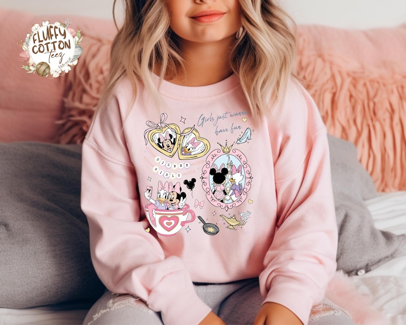 Puede incluir: Sudadera rosa claro con personajes de Disney, Minnie y Daisy. El dise&ntilde;o gr&aacute;fico incluye el texto "Girls just wanna have fun". El dise&ntilde;o incorpora corazones, coronas y otros elementos decorativos.