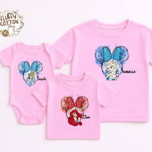 以下が含まれることがあります： ピンク色のベビーボディスーツとTシャツ。ディズニーのプリンセスデザインが特徴です。シンデレラ、エルサ、アリエルが描かれ、それぞれリボンと名前が付いています。家族やグループに最適です。
