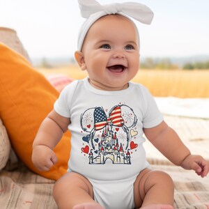 May include: A white baby onesie featuring a patriotic design. The graphic includes Mickey Mouse ears with the American flag, a castle, hearts, and fireworks. The baby is wearing a white headband and is smiling.