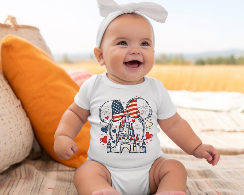 May include: A white baby onesie featuring a patriotic design. The graphic includes Mickey Mouse ears with the American flag, a castle, hearts, and fireworks. The baby is wearing a white headband and is smiling.