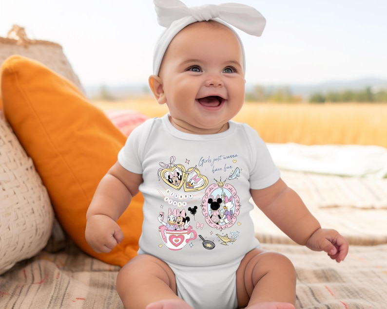 Puede incluir: Un beb&eacute; sonriente con un body blanco con gr&aacute;ficos de dibujos animados coloridos de Minnie y Daisy. El texto "Girls just wanna have fun" est&aacute; impreso en el body. El beb&eacute; tambi&eacute;n lleva una diadema blanca.
