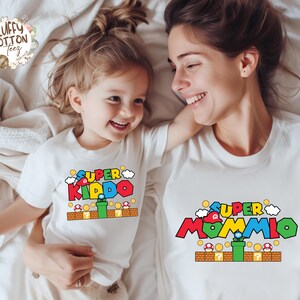 May include: Two white t-shirts with colorful graphic designs. One shirt says "SUPER KIDDO" and the other says "SUPER MOMMIO", both with a video game theme. The shirts are on a white bedspread.