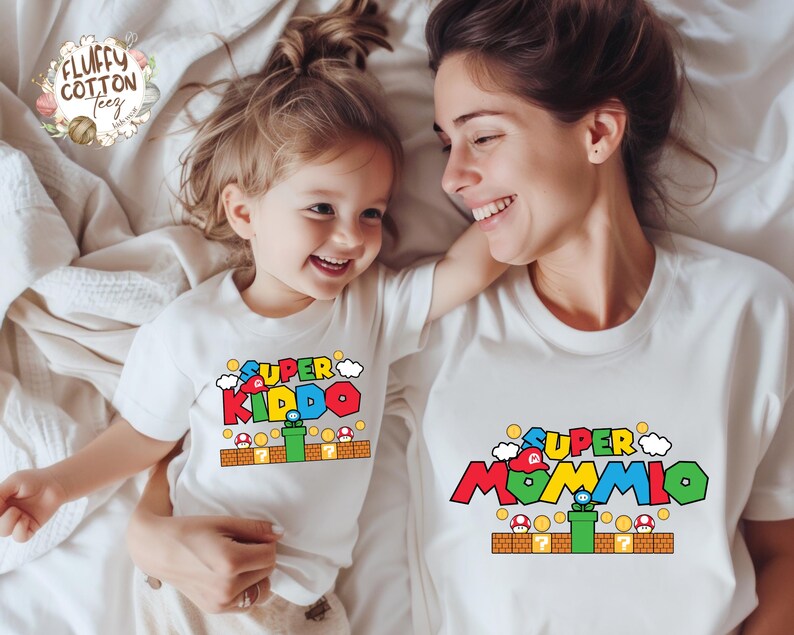 May include: Two white t-shirts with colorful graphic designs. One shirt says "SUPER KIDDO" and the other says "SUPER MOMMIO", both with a video game theme. The shirts are on a white bedspread.