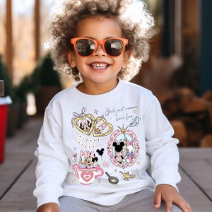 Puede incluir: Una sudadera blanca de manga larga con los personajes de dibujos animados Minnie Mouse y Daisy Duck. El dise&ntilde;o incluye corazones, una taza de t&eacute; y la frase "Girls just wanna have fun." El ni&ntilde;o lleva gafas de sol naranjas.