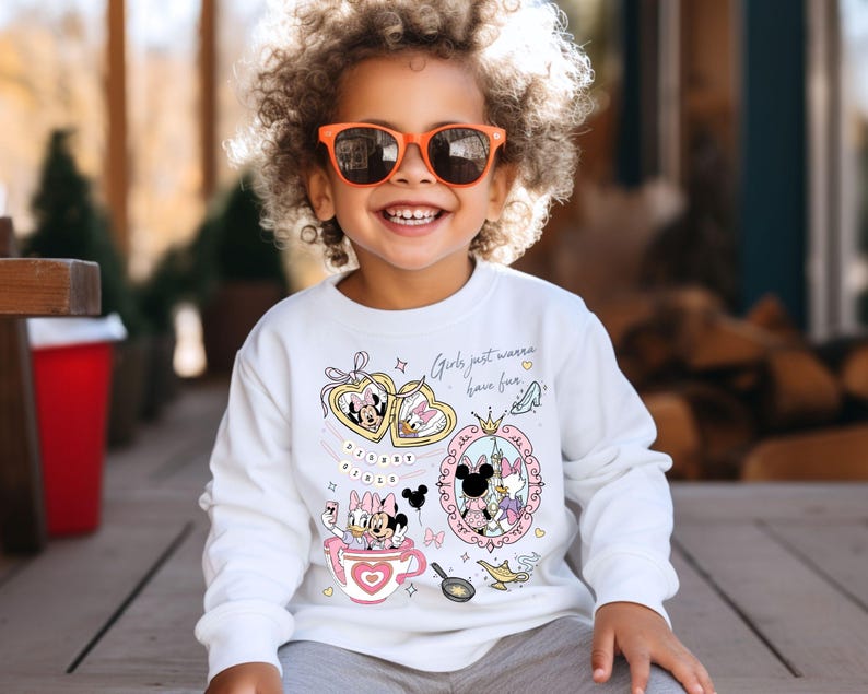 Puede incluir: Una sudadera blanca de manga larga con los personajes de dibujos animados Minnie Mouse y Daisy Duck. El dise&ntilde;o incluye corazones, una taza de t&eacute; y la frase "Girls just wanna have fun." El ni&ntilde;o lleva gafas de sol naranjas.