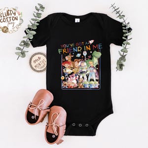Op de afbeelding: Zwarte babybody met een kleurrijke afbeelding van personages uit de film "Toy Story" en de tekst "You've got a friend in me". De body wordt getoond met bruine babyschoenen.