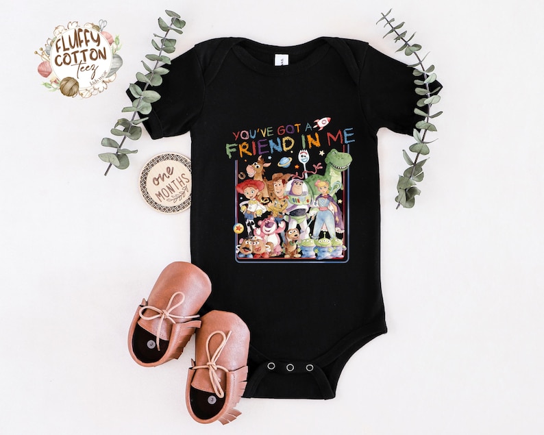 Op de afbeelding: Zwarte babybody met een kleurrijke afbeelding van personages uit de film "Toy Story" en de tekst "You've got a friend in me". De body wordt getoond met bruine babyschoenen.