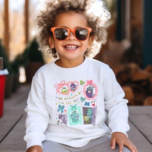 May include: A white long-sleeve sweatshirt featuring colorful cartoon character designs. The design includes framed images of monsters and the text "Monsters Inc.". The child is wearing orange sunglasses and smiling.