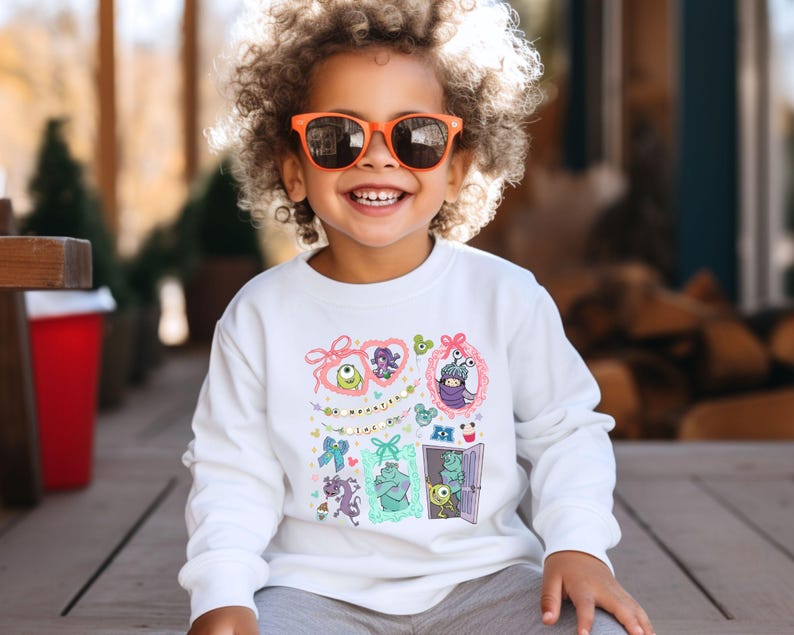 May include: A white long-sleeve sweatshirt featuring colorful cartoon character designs. The design includes framed images of monsters and the text "Monsters Inc.". The child is wearing orange sunglasses and smiling.