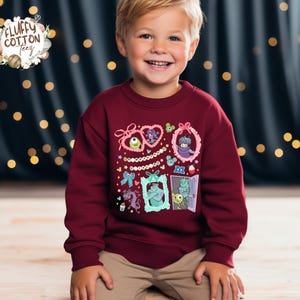 May include: A maroon sweatshirt featuring cartoon monster characters in decorative frames. The sweatshirt has a crew neck and long sleeves. The brand logo "Fluffy Cotton Teez" is in the upper left corner.