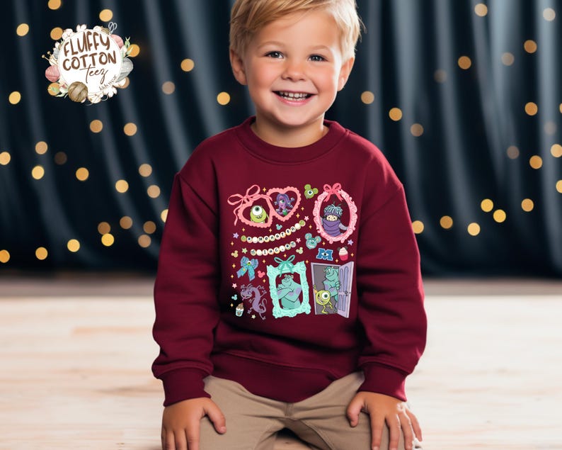 May include: A maroon sweatshirt featuring cartoon monster characters in decorative frames. The sweatshirt has a crew neck and long sleeves. The brand logo "Fluffy Cotton Teez" is in the upper left corner.
