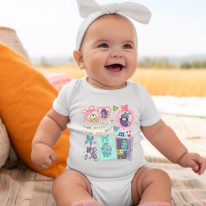 May include: A smiling baby wearing a white onesie with a colorful graphic featuring cartoon characters. The graphic includes various framed images of monsters and the text "& Boo". The baby is also wearing a white headband.