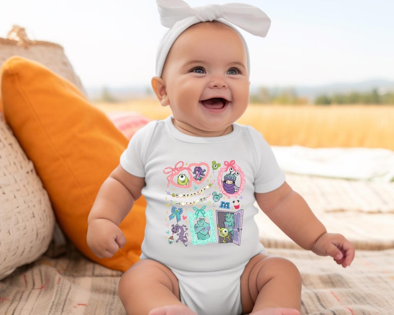 May include: A smiling baby wearing a white onesie with a colorful graphic featuring cartoon characters. The graphic includes various framed images of monsters and the text "& Boo". The baby is also wearing a white headband.