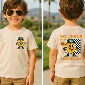 K&ouml;nnte beinhalten: Beiges T-Shirt mit einem Cartoon-Zitronen-Charakter, der eine gr&uuml;ne Kappe und schwarze Schuhe tr&auml;gt. Auf der Vorderseite des Shirts ist der Zitronen-Charakter zu sehen, w&auml;hrend auf der R&uuml;ckseite "My Jesus Makes Lemonade" mit einem Schachbrettmuster steht.