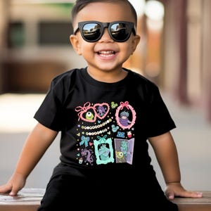 May include: Black t-shirt featuring colorful cartoon monster characters in decorative frames. The design includes pink bows, strings of pearls, and various monster illustrations. The shirt is worn by a child wearing black sunglasses.