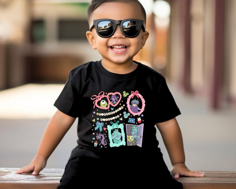 May include: Black t-shirt featuring colorful cartoon monster characters in decorative frames. The design includes pink bows, strings of pearls, and various monster illustrations. The shirt is worn by a child wearing black sunglasses.