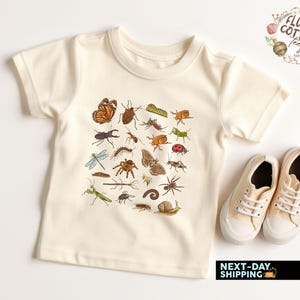 May include: Cream-colored t-shirt featuring a colorful insect illustration. The graphic includes a variety of bugs, such as butterflies, beetles, and spiders. The shirt is paired with a pair of beige canvas sneakers. The text "Fluffy Cotton Teez" is in the upper right corner.