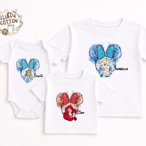 以下が含まれることがあります： ディズニーのプリンセスデザインが施された白いTシャツとボディスーツ。各衣服には、ミッキーマウスの耳の輪郭の中にプリンセスのイラストが描かれています。「Sophia」、「Isabella」、「Liam」という名前がプリントされています。