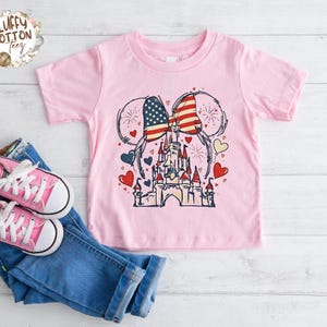May include: A light pink t-shirt featuring a graphic of a castle with American flag-themed mouse ears. The design includes red, white, and blue hearts and fireworks. Pink sneakers and blue jeans are next to the shirt.