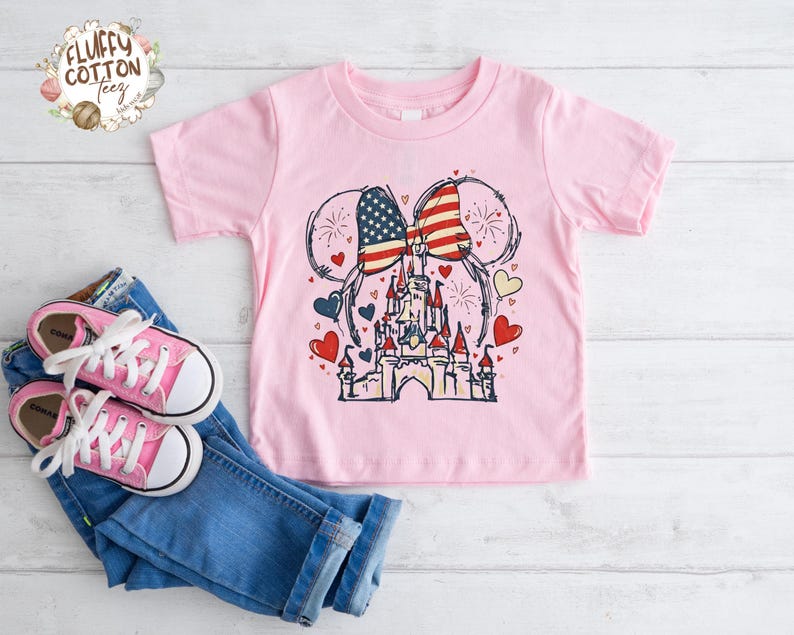May include: A light pink t-shirt featuring a graphic of a castle with American flag-themed mouse ears. The design includes red, white, and blue hearts and fireworks. Pink sneakers and blue jeans are next to the shirt.