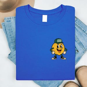 K&ouml;nnte beinhalten: K&ouml;nigsblaues T-Shirt mit einem Cartoon-Zitronen-Charakter, der eine gr&uuml;ne Kappe mit dem Text "JESUS SAVES" tr&auml;gt. Die Zitrone hat ein l&auml;chelndes Gesicht, schwarze Schuhe und wei&szlig;e Handschuhe. Das T-Shirt ist auf Jeans gefaltet.