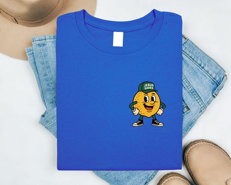 K&ouml;nnte beinhalten: K&ouml;nigsblaues T-Shirt mit einem Cartoon-Zitronen-Charakter, der eine gr&uuml;ne Kappe mit dem Text "JESUS SAVES" tr&auml;gt. Die Zitrone hat ein l&auml;chelndes Gesicht, schwarze Schuhe und wei&szlig;e Handschuhe. Das T-Shirt ist auf Jeans gefaltet.