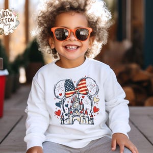 May include: A white long-sleeved sweatshirt featuring a patriotic design. The graphic includes a castle, hearts, and Mickey Mouse ears with an American flag pattern. The child is wearing orange sunglasses and smiling.