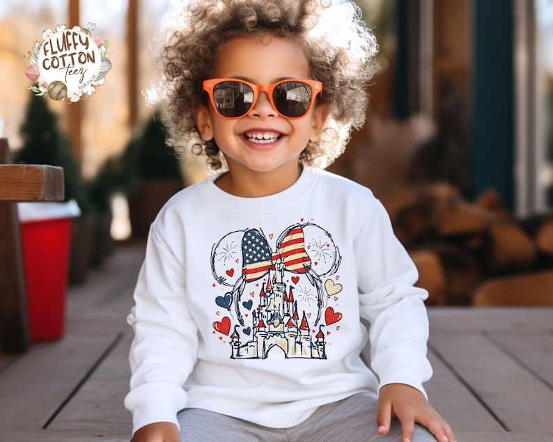 May include: A white long-sleeved sweatshirt featuring a patriotic design. The graphic includes a castle, hearts, and Mickey Mouse ears with an American flag pattern. The child is wearing orange sunglasses and smiling.