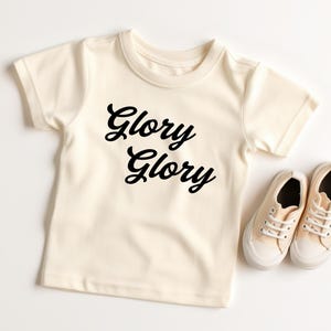 May include: A cream colored short-sleeved t-shirt with the words "Glory Glory" printed in black on the front.