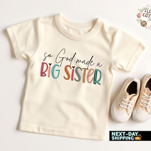 May include: A white short-sleeved t-shirt with the text "So God made a BIG SISTER" in colorful letters. The text is in a playful font with hearts.