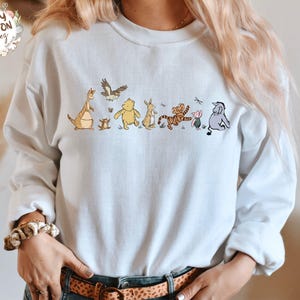 May include: A light blue sweatshirt with a graphic of Winnie the Pooh and his friends. The characters are in a line, walking from left to right. The characters include Tigger, Piglet, Eeyore, Kanga, Roo, and Rabbit.