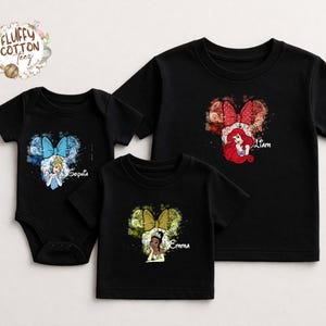 以下が含まれることがあります： 黒いTシャツと、ディズニーのプリンセスデザインのボディスーツ。シンデレラ、アリエル、ティアナのデザインで、それぞれミッキーマウスの耳の形をした水彩画のスプラッシュの中に描かれています。キャラクターの下には名前が印刷されています。「Fluffy Cotton Teez」のブランドロゴが左上にあります。