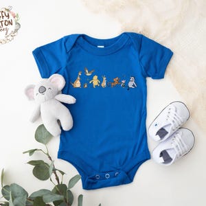 May include: A royal blue baby onesie with a graphic of characters from the movie 'The Lion King'. The characters include Simba, Timon, Pumbaa, and Zazu. The onesie has short sleeves and a snap closure.