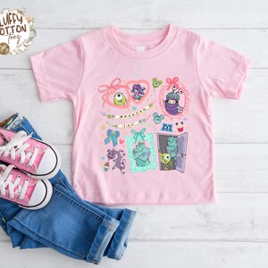 May include: A light pink t-shirt featuring a colorful graphic of characters from the movie "Monsters, Inc." The design includes framed images of the monsters, bows, and the text "Monster Collection". Pink canvas sneakers and blue jeans are also in the image.