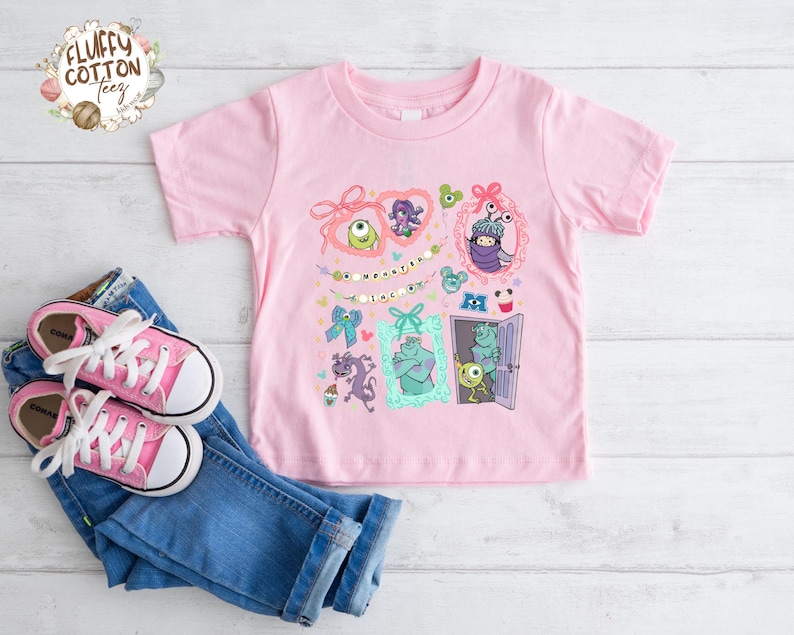 May include: A light pink t-shirt featuring a colorful graphic of characters from the movie "Monsters, Inc." The design includes framed images of the monsters, bows, and the text "Monster Collection". Pink canvas sneakers and blue jeans are also in the image.