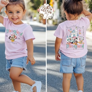 May include: A light pink t-shirt with a Disneyland graphic, worn with denim shorts and white sneakers. The front of the shirt features the Disneyland logo and characters. The back of the shirt shows a castle and more characters.
