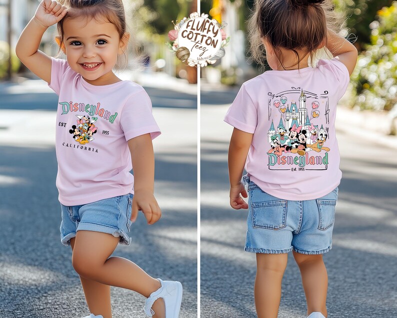 May include: A light pink t-shirt with a Disneyland graphic, worn with denim shorts and white sneakers. The front of the shirt features the Disneyland logo and characters. The back of the shirt shows a castle and more characters.
