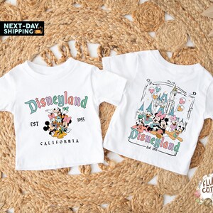 May include: Two white t-shirts with Disneyland graphics. One shirt features the Disneyland logo with Mickey Mouse and friends, the text "EST 1955 CALIFORNIA". The other shirt has a castle graphic with the Disneyland logo and the text "Est. 1955".