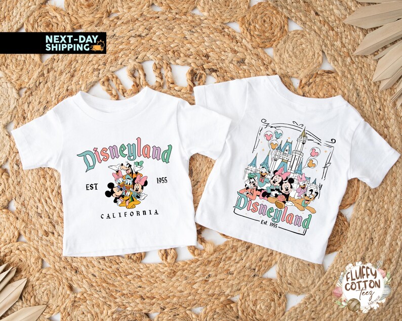 May include: Two white t-shirts with Disneyland graphics. One shirt features the Disneyland logo with Mickey Mouse and friends, the text "EST 1955 CALIFORNIA". The other shirt has a castle graphic with the Disneyland logo and the text "Est. 1955".