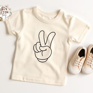 May include: Cream-colored toddler t-shirt featuring a black cartoon hand making a peace sign. The shirt is paired with matching beige canvas sneakers with white laces. The Fluffy Cotton Teez logo is in the upper left corner.