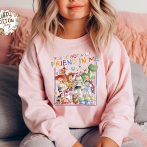 Op de afbeelding: Een lichtroze sweatshirt met een kleurrijke afbeelding van personages uit de film "Toy Story". De tekst "You've Got a Friend in Me" staat boven de afbeelding. De sweatshirt heeft lange mouwen en een ronde hals.