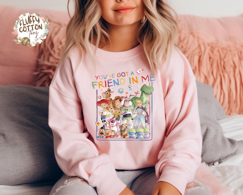Op de afbeelding: Een lichtroze sweatshirt met een kleurrijke afbeelding van personages uit de film "Toy Story". De tekst "You've Got a Friend in Me" staat boven de afbeelding. De sweatshirt heeft lange mouwen en een ronde hals.