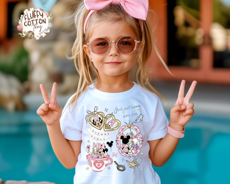 Puede incluir: Una camiseta blanca con gr&aacute;ficos coloridos de personajes de Disney, incluyendo a Minnie y Daisy, y el texto "Girls just wanna have fun." La camiseta la lleva una persona joven con una diadema rosa y gafas de sol.