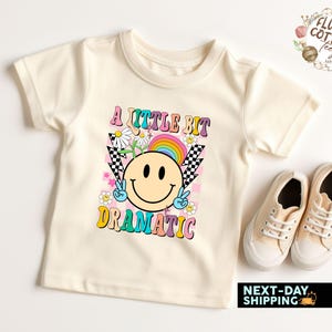 May include: A white t-shirt with a colorful graphic design featuring a smiley face, flowers, and the text "A little bit dramatic".